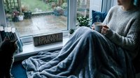 Young Caucasian woman wrapped in soft gray blanket sitting by rainy window with hot drink, cozy stay home hygge lifestyle concept.