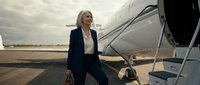 Middle-aged woman with blonde hair, Caucasian ethnicity, wearing dark business suit and cream blouse, boarding private jet on airport tarmac during daytime. Suitable for corporate travel, business lifestyle, executive transportation, and professional aviation concepts.