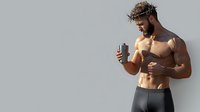 Muscular shirtless Caucasian male athlete with beard holding protein shake after workout, wearing black shorts against light gray background. Fitness lifestyle concept.