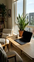 Modern home office workspace with laptop, snake plant and coffee cup by sunny window overlooking urban buildings for remote work productivity.