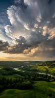 Dramatic mammatus clouds loom over rolling green hills and winding river valley at golden hour, showcasing nature's atmospheric power and scenic landscape beauty.
