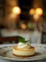 Classic cheesecake slice on white plate garnished with whipped cream and fresh mint leaf, soft bokeh lights creating romantic restaurant atmosphere.