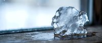 Clear ice chunk melting on wooden surface with soft natural lighting and blurred background for refreshing beverage and cooling concepts.