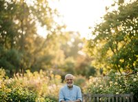 Senior man with glasses wearing light blue shirt sitting on wooden bench in lush garden surrounded by green foliage and flowering plants during golden hour sunlight, outdoor portrait for lifestyle and wellness content.