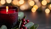 Red glass candle holder with burning flame surrounded by holly berries and eucalyptus leaves against soft golden bokeh lights creating cozy holiday atmosphere.