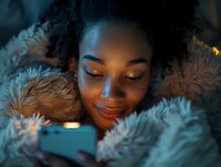 Young African American woman smiling while using smartphone wrapped in cozy faux fur blanket, warm evening lighting creates intimate atmosphere.