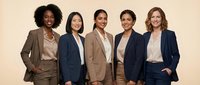 Diverse group of five professional women wearing business blazers and casual tops standing together against neutral beige background for corporate team, leadership, and workplace diversity concepts.