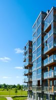 Modern residential apartment building with glass balconies and metal railings overlooking green landscape under clear blue sky. Suitable for real estate, architecture, and urban development projects.