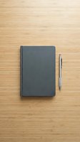 Closed dark gray notebook with silver pen on light bamboo wood surface, flat lay composition for business, education, planning, writing, and office workspace design projects.