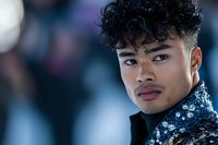 Young Asian male model with curly hair wearing sequined jacket poses against blurred bokeh background in dramatic lighting and modern fashion style.