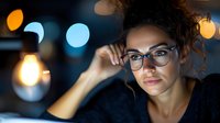 Young woman with glasses working late at night, illuminated by soft light with bokeh background, conveying focus and determination.