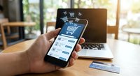 Male hand holding smartphone with online shopping app interface, shopping cart icons floating above screen, laptop and credit card on wooden desk in bright office.