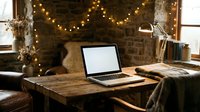Laptop with blank screen on rustic wooden desk decorated with warm string lights, cozy blankets, and dried flowers for remote work or creative projects.