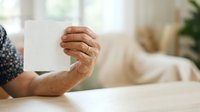 Senior adult with brown skin tone holding blank white card or paper at wooden table indoors, suitable for greeting cards, announcements, messages, and promotional campaigns.