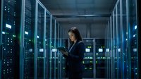 Female IT specialist working with tablet in modern data center server room with blue LED lighting and network equipment racks for technology solutions.