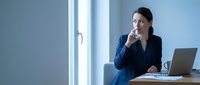 Thoughtful female professional in blue blazer sitting at desk with laptop, hand near chin in contemplative pose by bright window, representing business strategy and decision-making.