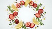 Fresh ingredients wreath with sliced tomatoes, lemon wedges, rosemary sprigs and peppercorns arranged in circle on white background for culinary design.