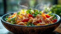 Steaming hot Asian noodle bowl with colorful vegetables, shredded chicken, fresh herbs and garnish served in rustic ceramic dish on light surface.