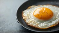 Close-up of sunny side up fried egg with crispy edges and bright orange yolk on black plate against dark background, seasoned with black pepper.