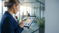 Caucasian businesswoman in navy blazer using magnifying glass over tablet with employee profiles in modern office.