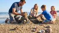 Adult male with reddish-brown hair and fair complexion, wearing navy shirt, builds sandcastle with three children on sandy beach near ocean waves on sunny day. Family leisure activity and summer vacation concept.