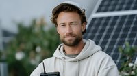 Young Caucasian man with beard wearing gray hoodie smiling near solar panels, representing renewable energy and sustainable lifestyle choices.