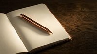 Open notebook with copper pen on rustic wooden desk surface for writing journaling planning and creative work projects.