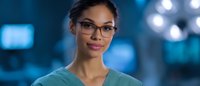 Mixed race female medical professional wearing glasses and scrubs smiling confidently in hospital setting with blurred blue background and bokeh lights.