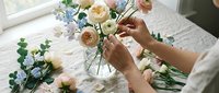 Female florist hands arranging fresh roses and eucalyptus in glass vase on white table. Professional flower arrangement workshop for wedding bouquet creation.