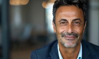 Confident mature Mediterranean businessman with salt and pepper hair smiling warmly at camera, wearing navy blazer against blurred office background.
