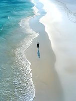 Solitary figure walking along pristine white sand beach with turquoise ocean waves creating serene coastal landscape from aerial perspective.