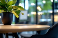 Green plant in black pot on wooden round table with blurred modern office or cafe interior background, creating cozy minimalist workspace atmosphere.