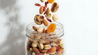 Mixed nuts and dried fruits falling into glass jar on white background, suitable for food packaging, healthy snack marketing, and nutrition content.