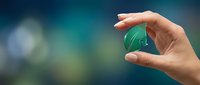 Woman's hand holding small green leaf-shaped object against blurred teal bokeh background, symbolizing eco concept and sustainability.