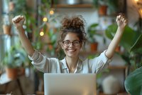 Young mixed race woman with glasses celebrating success at laptop, arms raised in victory gesture among houseplants in cozy home office setting with bokeh lights.