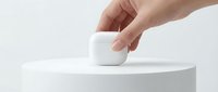 Hand placing a white wireless earbuds charging case on a minimalist white podium, clean studio background, modern tech lifestyle concept.