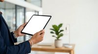 Businessman in dark blue suit holding tablet with blank screen in modern office space with potted plant and natural light.