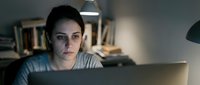 Young woman working late at night on laptop computer in dark office environment with focused concentration and desk lamp illumination for productivity concepts.