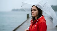 Young woman with long dark hair and fair complexion wearing a bright red jacket, holding a transparent umbrella while standing on a wooden pier during rainy weather with blurred water and overcast sky in background.