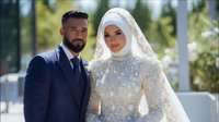 Middle Eastern couple on wedding day, bride in elegant white hijab and embellished dress, groom in navy suit, standing together in outdoor setting with soft natural light.