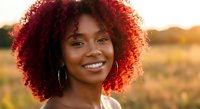 Young African American woman with curly red hair smiling warmly at golden hour sunset. Natural beauty portrait with soft lighting for lifestyle content.