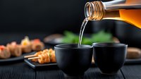 Pouring sake from bottle into traditional black ceramic cups with sushi rolls in background, dramatic lighting creates atmospheric dining scene.