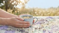 Hands holding ceramic mug with herbal tea on lavender field during golden hour sunset creating peaceful morning ritual atmosphere.