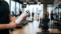 Fit man holding water bottle in modern gym with equipment and blurred people exercising in background, hydration during workout.