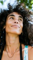 Young woman with dark curly hair and freckles looking upward outdoors, wearing gold necklace and hoop earrings, ideal for lifestyle, beauty, and wellness marketing campaigns.