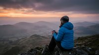 Young adult female in blue winter jacket and beanie sitting on mountain peak at sunrise, overlooking misty valleys and rolling hills. Outdoor adventure and hiking lifestyle photography.