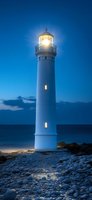 White lighthouse illuminated at dusk with bright beacon light against deep blue sky and ocean, rocky shore in foreground creating dramatic coastal scene.