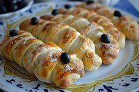 Freshly baked puff pastry rolls filled with cream cheese, topped with black olives and golden crust, served on decorative plate for appetizer or party snack.