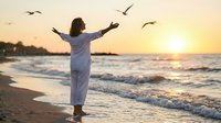 Woman in white clothing with arms raised celebrating freedom on beach at sunset with seagulls flying overhead. Peaceful ocean waves and golden hour lighting.
