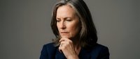 Middle-aged woman with shoulder-length dark hair and fair complexion wearing navy blazer, hand near chin in thoughtful pose against neutral gray background for business and professional concepts.